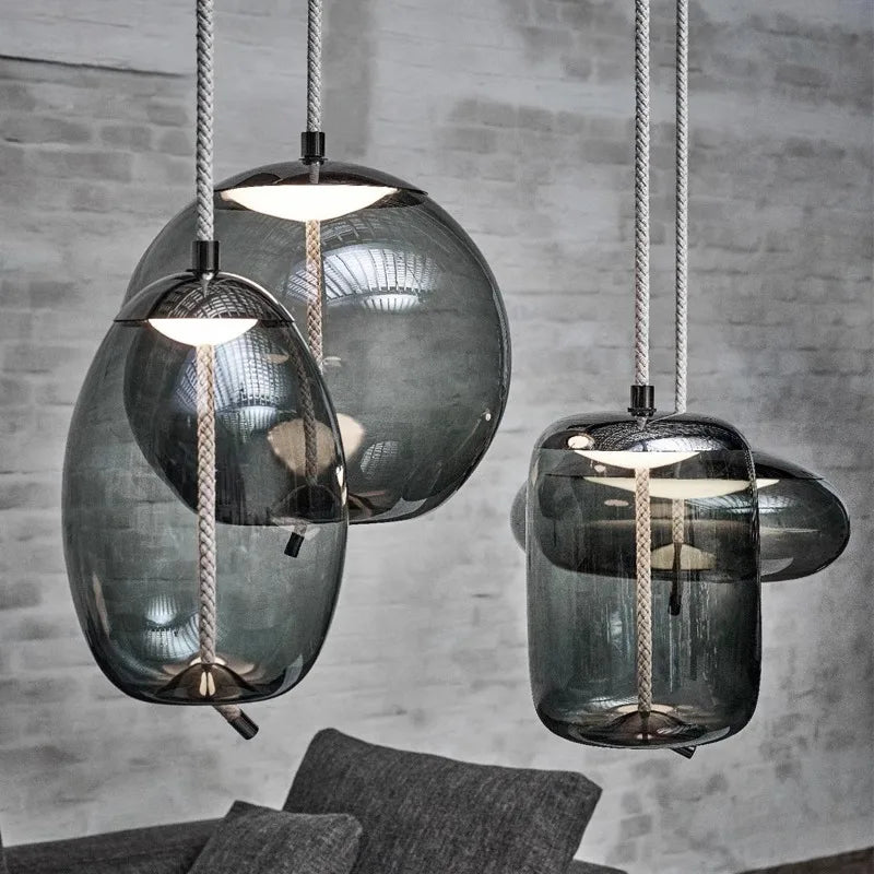 Three smoked glass pendant lights with textured cords hanging above a gray sofa against a concrete wall, showcasing the Modern LED Pendant Light — Chandelier for Living & Dining Room in a minimalist setting.