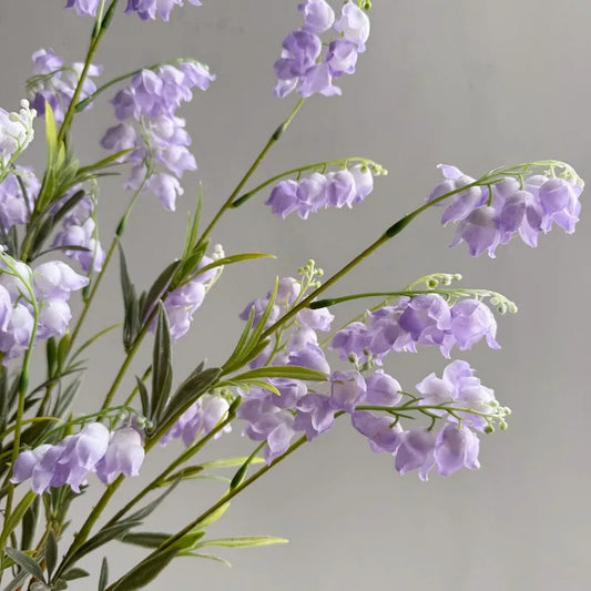 Lavender artificial lily of the valley stems with green leaves against soft gray background