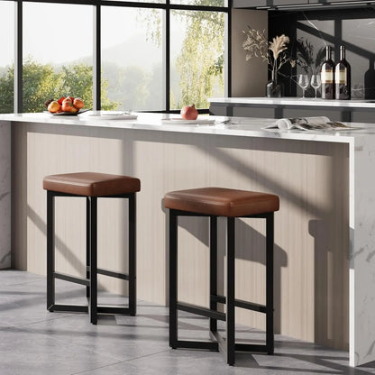 Two modern 24" Bar Stools Set of 2 — Backless Counter Height with PU Leather Seat in brown with black metal legs positioned in a bright kitchen with a marble countertop and large windows.
