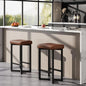 Two modern 24" Bar Stools Set of 2 — Backless Counter Height with PU Leather Seat in brown with black metal legs positioned in a bright kitchen with a marble countertop and large windows.