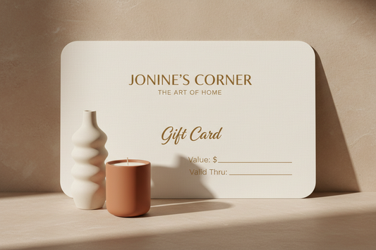 Jonine's Corner gift card displayed alongside a minimalist white sculptural vase and a terracotta-colored candle, set against a warm neutral background.